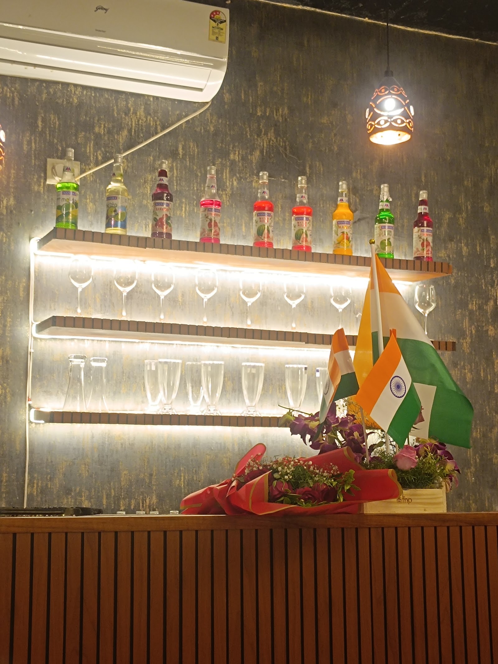 The bar back — a row of fruit syrup bottles above two lit shelves of wine glasses and tall milkshake glasses, against a textured grey and gold wall
