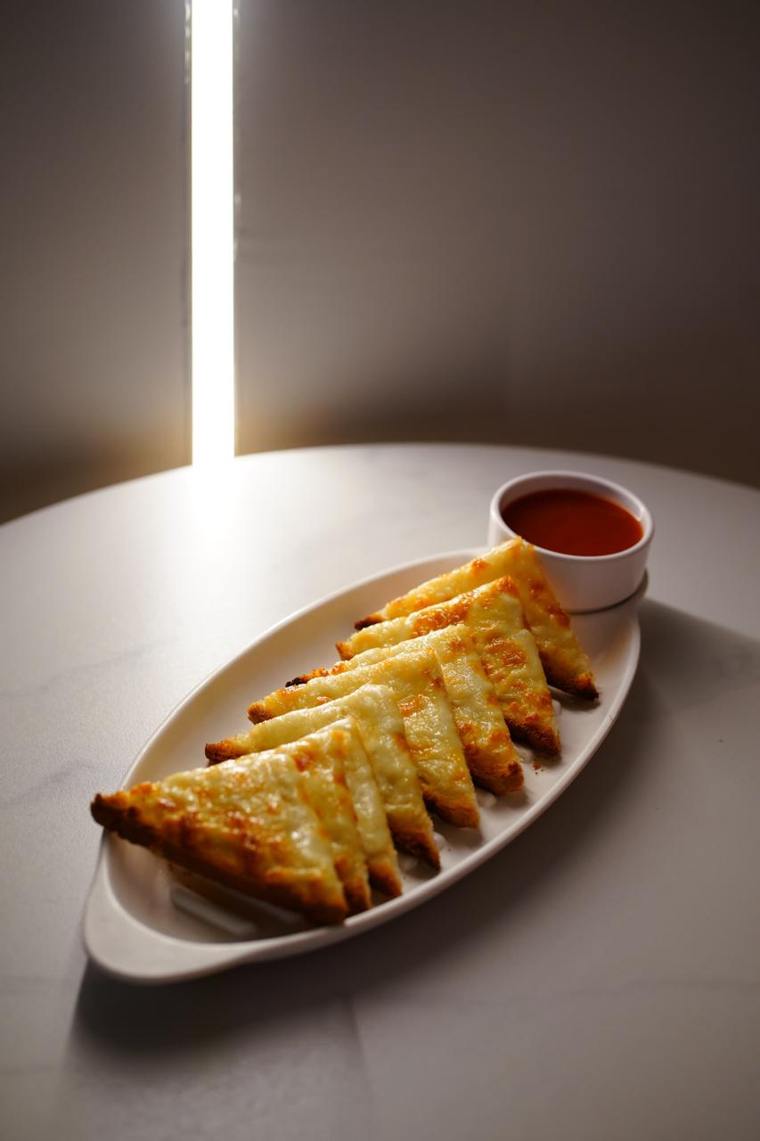 Cheese garlic bread cut into six triangles on a white oval plate, grilled until the cheese browns, with a pot of red dipping sauce