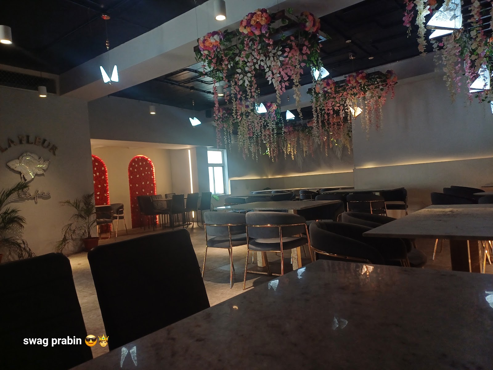 The main room at La Fleur — the illuminated LA FLEUR Cafe rose sign on a pale wall, two red flower arches behind it, a canopy of pink and white hanging blossom under a black industrial ceiling, and marble tables with grey velvet chairs on gold legs