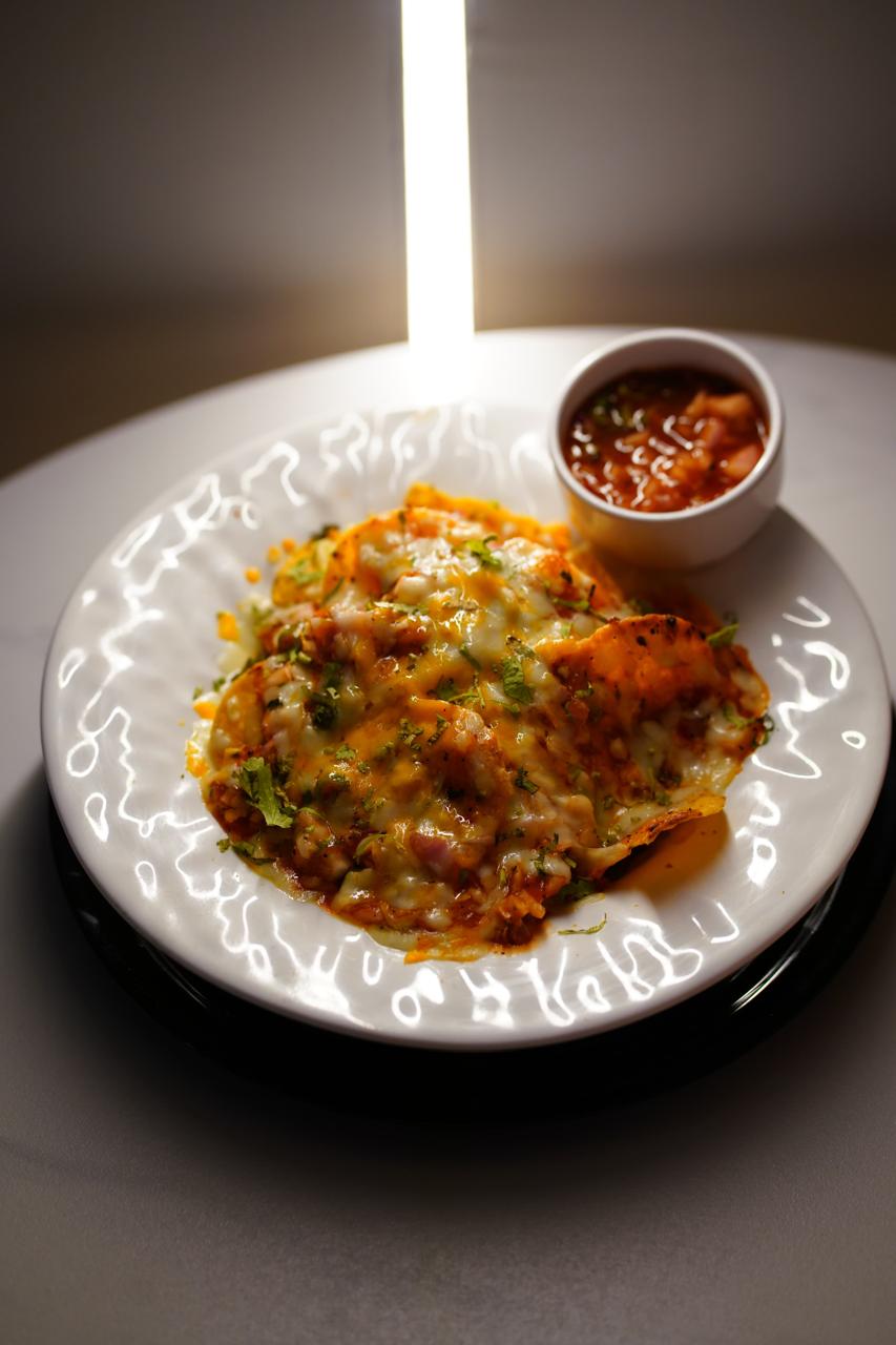 Overload nachos on a patterned white plate — tortilla chips under melted cheese, salsa and chopped coriander, with a pot of salsa alongside
