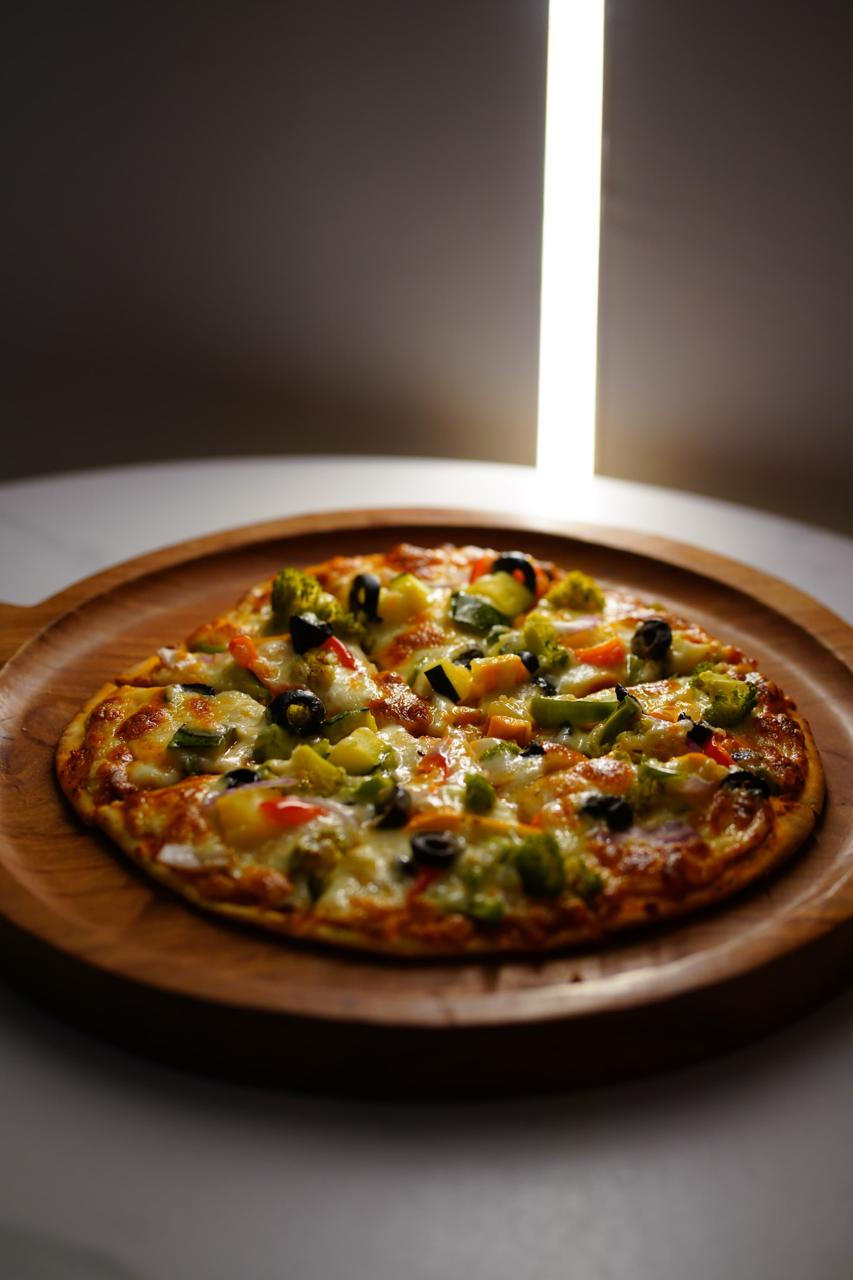 A vegetable pizza on a round wooden board — broccoli, black olives, peppers, sweetcorn and red onion on melted mozzarella