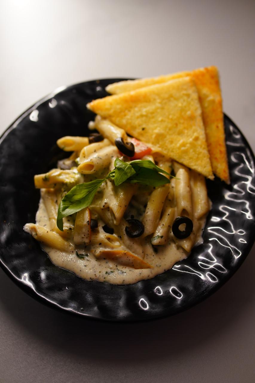 Penne alfredo in a black ceramic bowl — cream sauce with black olives, peppers and fresh basil, with two triangles of garlic bread standing in it