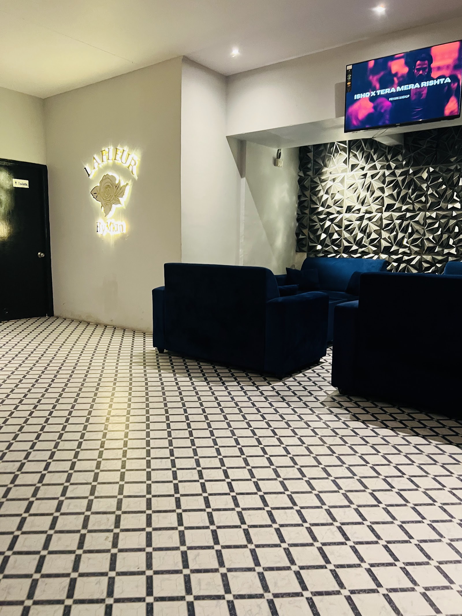 The lounge — a gold illuminated LA FLEUR rose crest lettered 'By Mun' on a cream wall, navy velvet sofas against a faceted black-and-white feature wall, and a black-and-white diamond tiled floor