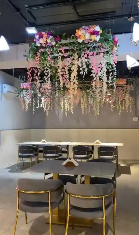 A hanging floral canopy of peonies and trailing pink and white blossom suspended over marble-topped tables and grey velvet chairs with gold frames