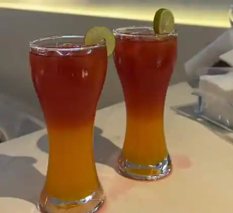 Two tall mocktail glasses on a counter, each graduating from orange at the base to deep red at the top, finished with a slice of lime